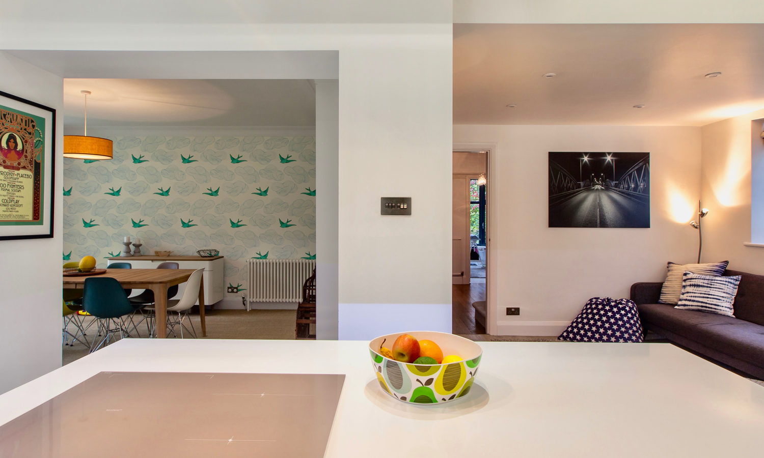 View from modern open plan white kitchen island looking into dining area with bird wallpaper and sofa seating space with grey sofa