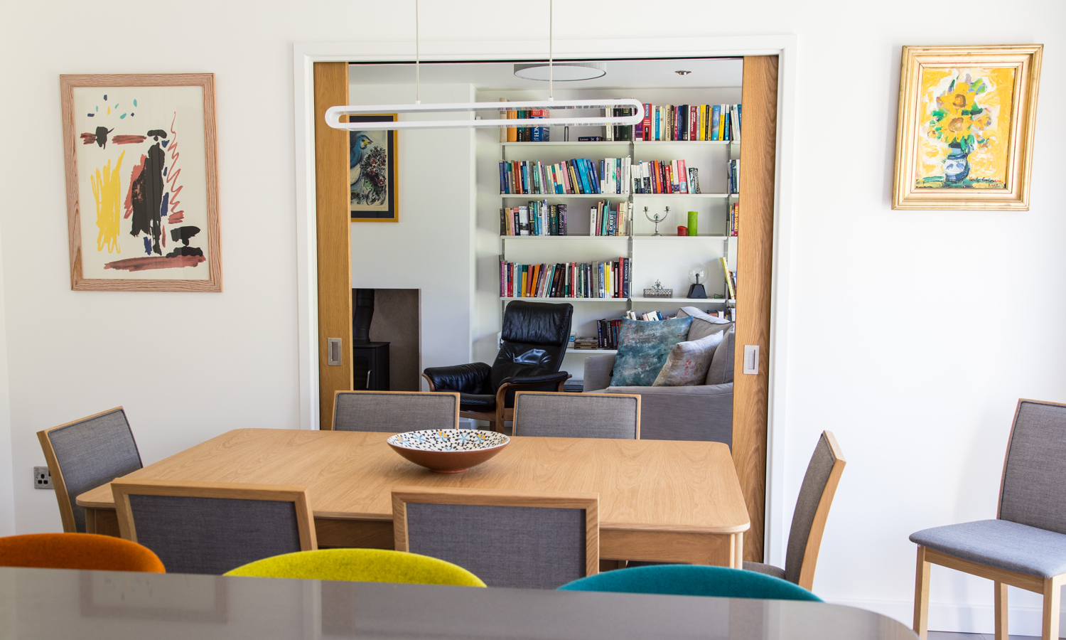 Modern family dining table with linear light over, pocket doors into living room space, oak table, white walls, colourful artwork.