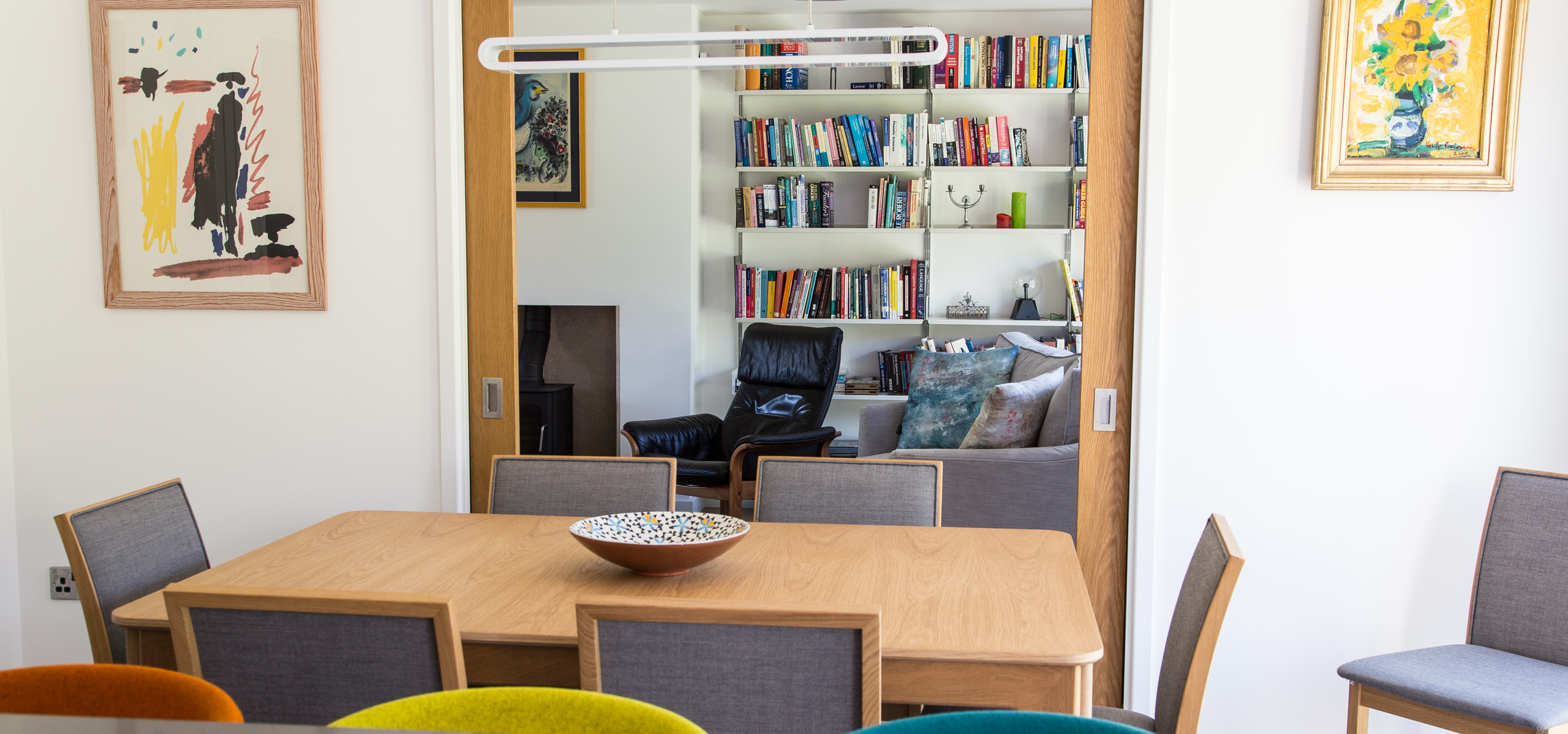 Modern family dining table with pocket doors into living room space. Oak table and doors and white walls, with colourful artwork and stools.